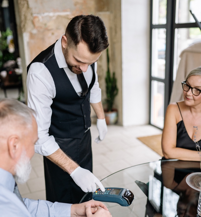 Waiter Using a Terminal at a Table where an Elderly Couple Sits